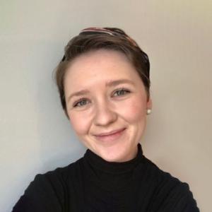 A portrait of Charlotte, an Eli's Place Peer Advisor. She has short, styled hair held back by a headband and is wearing a black high-neck top. She is smiling warmly at the camera against a plain, neutral background.