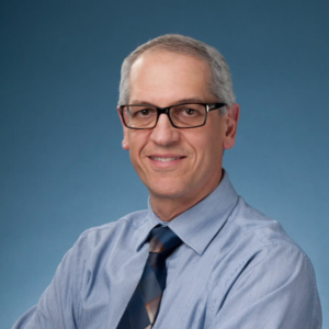 A professional headshot of Dr. Howard Hamer, smiling warmly at the camera. He has short, graying hair and is wearing dark-rimmed glasses, a light blue dress shirt, and a dark, patterned tie, set against a plain, light blue background.