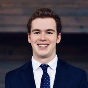 A professional headshot of a young man with short, wavy brown hair, smiling brightly at the camera. He is wearing a dark blue suit jacket over a white dress shirt and a dark blue tie with small white polka dots, set against a dark, out-of-focus background.