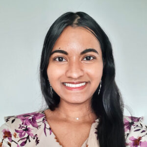 A portrait of Lucksini Raveendran, an Eli's Place Peer Advisor. She has long, dark hair, is wearing a white top with a purple and black floral pattern and silver dangling earrings, and is smiling warmly at the camera against a neutral, light-gray background.