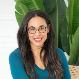 A professional headshot of Deanne Simms, smiling warmly at the camera. She has long, dark wavy hair, is wearing dark-rimmed glasses and a teal-colored long-sleeved top, set against a background featuring large, lush green tropical leaves.