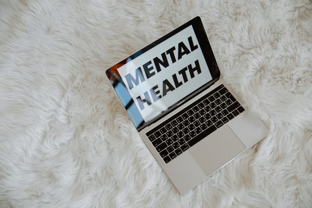 An overhead shot of an open laptop computer displaying the words "MENTAL HEALTH" in large, bold letters on its screen, resting on a fluffy white shag rug or faux fur surface.