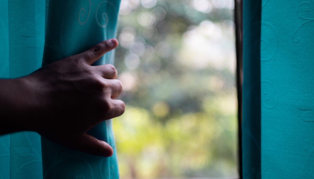 A close-up photograph capturing a hand drawing back a heavy, dark-colored curtain to let light stream into a dimly lit room through a large glass window.