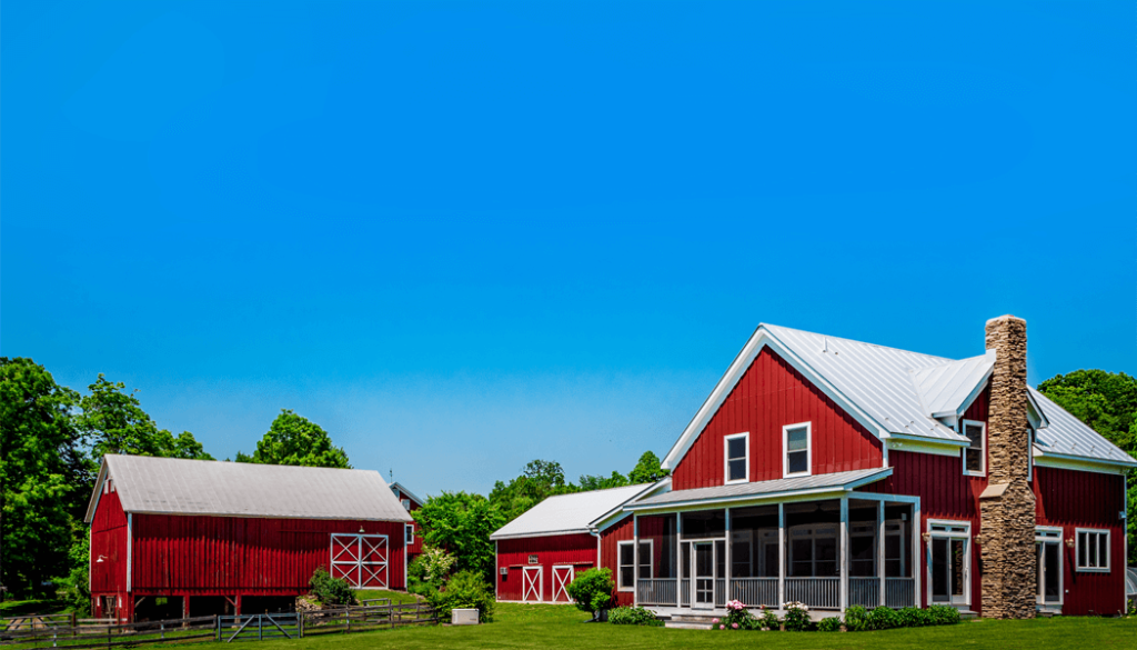 A scenic photograph of a rustic two-story red farmhouse with a white steel roof and a prominent stone chimney on the right. To the left, a large matching red barn stands behind a wooden fence. The buildings are set on a lush green lawn under a clear, bright blue sky.