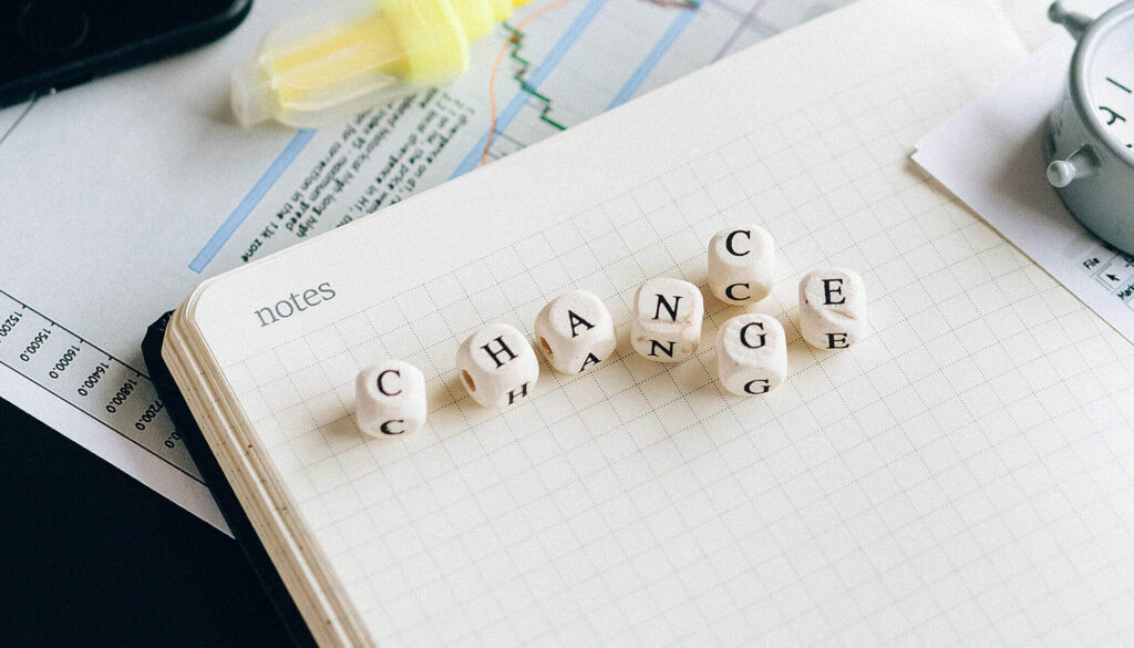 A top-down photograph of an open notebook with the word "notes" printed on the left page. Small wooden letter cubes are scattered across the pages, spelling out the word "CHANGE." In the softly blurred background, a pen, a clock, and document papers with financial charts are visible.