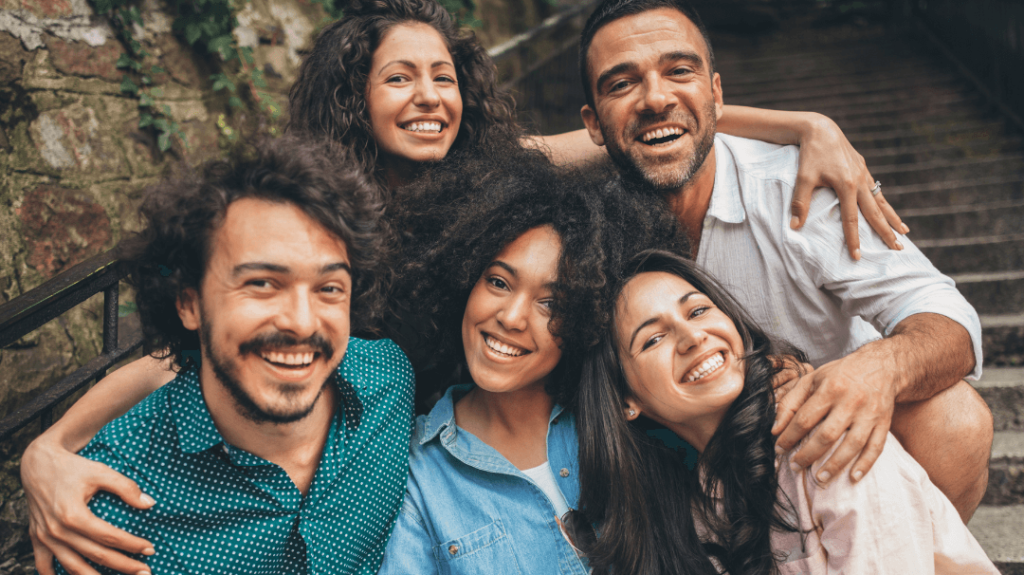 A close-up, joyful group portrait of five diverse adults smiling and hugging tightly on an outdoor stone staircase. The group includes three women and two men with expressive, happy faces.