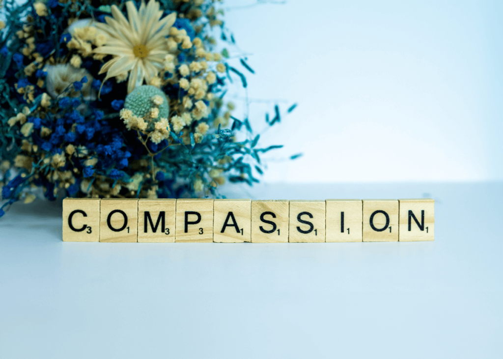 A conceptual close-up photograph featuring Scrabble-style wooden tiles laid out on a white surface to spell the word "COMPASSION". The tiles are positioned in a straight line at the bottom of the frame, with the letters clearly visible: C, O, M, P, A, S, S, I, O, N. In the upper-left corner, a soft-focus, blurred floral arrangement of dried yellow, blue, and cream-colored flowers adds a gentle, aesthetic touch.