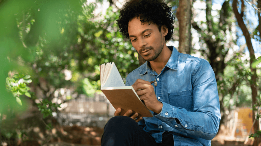 A close-up photograph of a man with dark curly hair and a light denim button-up shirt sitting outside on a sunny day. He is focused on writing in a leather-bound notebook with a pen, surrounded by soft, blurred green tree leaves in the foreground and a sun-dappled wooded background.