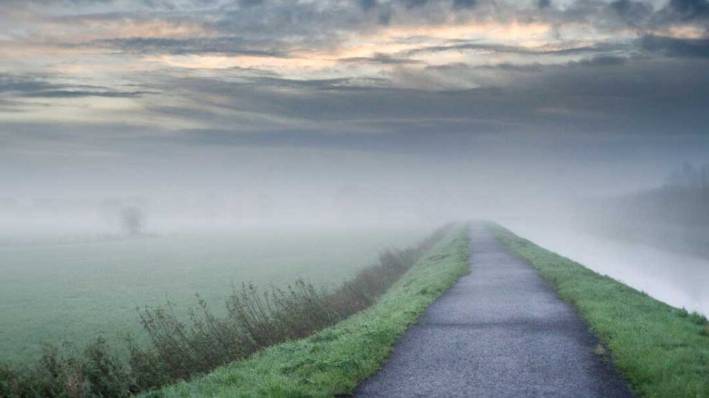 A scenic, wide-angle photograph of a narrow asphalt pathway cutting straight through a foggy green field under a dramatic, overcast sky. Dense mist blankets both sides of the path, obscuring the distant horizon and creating a soft, quiet atmosphere.