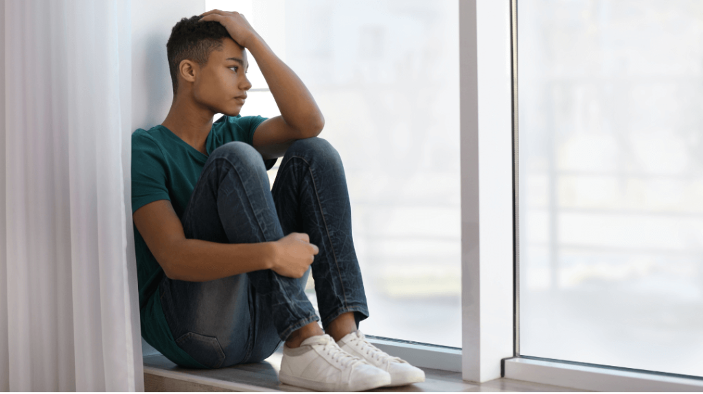 A poignant, full-shot photograph of a young person sitting on a wide white windowsill inside a bright, sunlit room. They are wearing a dark teal t-shirt, blue jeans, and white sneakers, sitting with their knees pulled up tightly to their chest. One hand is resting heavily on their forehead as they gaze thoughtfully and sadly out the large window, looking toward a blurred outdoor backdrop.
