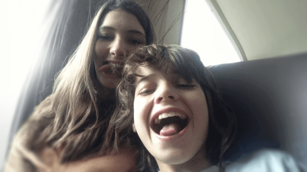 A grainy, nostalgic close-up selfie of two young siblings sharing a playful moment inside a vehicle. In the foreground, a young boy with medium-length dark hair smiles widely with his mouth open in excitement. Just behind him, an older girl with long brown hair looks into the camera, playfully sticking her tongue out.