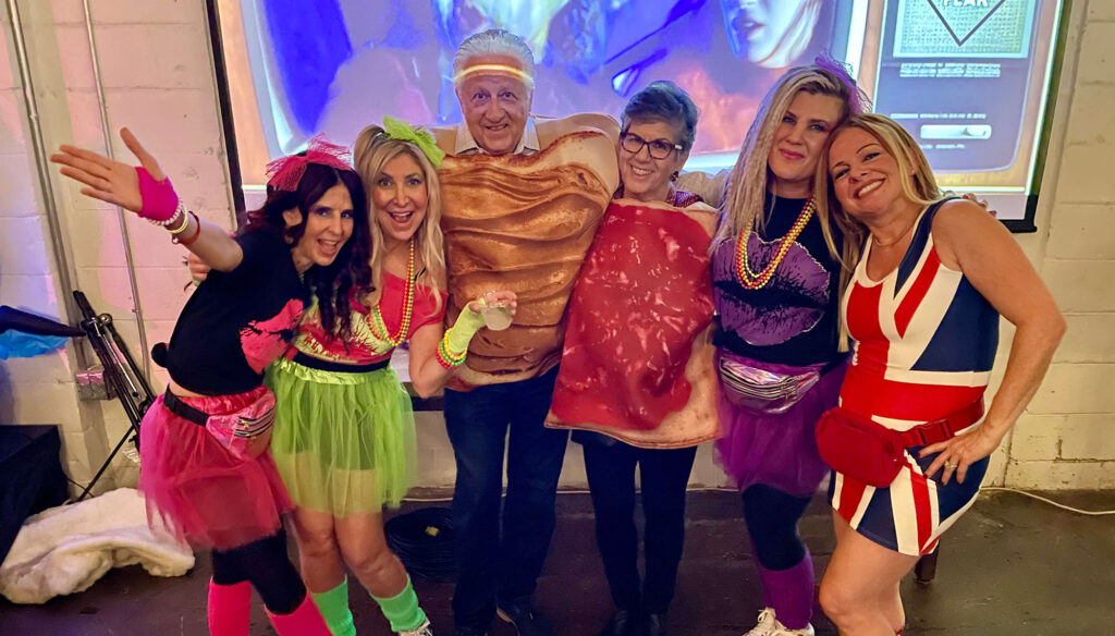 A group of six smiling people pose together in vibrant retro costumes at an indoor social event under blue lighting. The group includes a man in an orange ruffled top with neon glow stick headbands, a woman in a British Union Jack flag dress with a red fanny pack, a woman in a purple and neon green tiered tutu skirt, and others sporting colorful bead necklaces and bright accessories.
