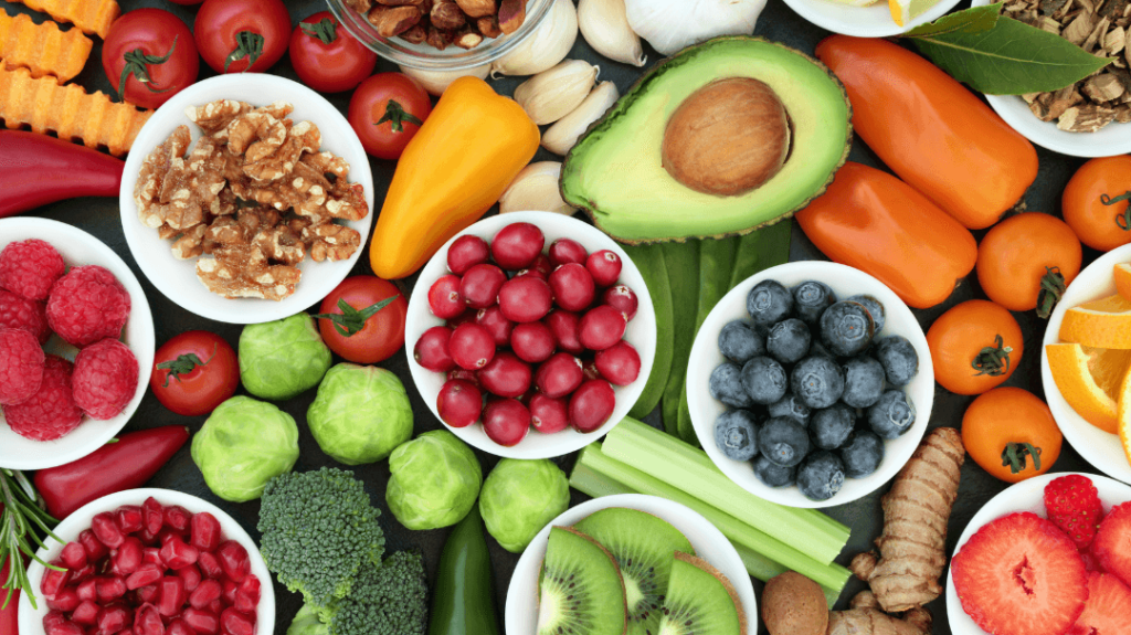 A vibrant, overhead close-up shot of a dark surface filled with small white bowls containing fresh, colorful foods. The bowls are neatly arranged with red raspberries, green Brussels sprouts, bright red pomegranate seeds, chopped walnuts, fresh cranberries, and blueberries. Surrounding the bowls are scattered whole ingredients including an open avocado, crisp celery sticks, sliced kiwi, fresh ginger, a whole strawberry slice, and orange wedges, creating a diverse display of healthy nutrition.