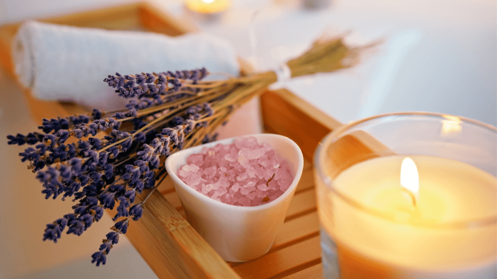 A peaceful wellness ritual featuring a bundle of dried lavender, a small heart-shaped bowl filled with pink bath salts, and a lit candle on a wooden bath tray.
