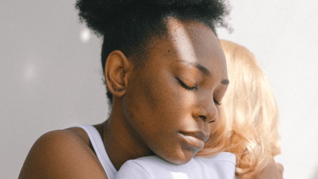 A close-up photograph of a Black individual with their eyes closed, receiving a comforting embrace from a blonde person whose face is partially obscured.