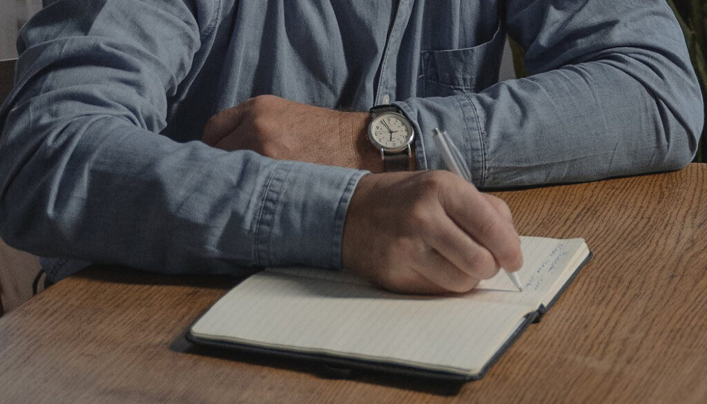A close-up of a person's hands holding a green pen, writing thoughtfully in a blank, open notebook.