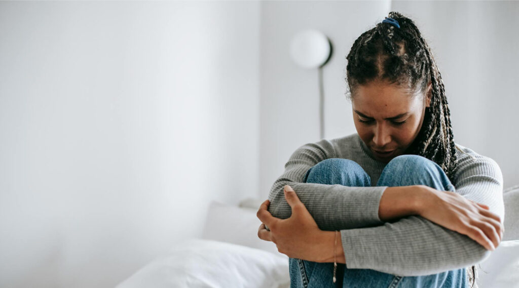 A person with braided hair sitting down and tightly hugging their knees to their chest, looking down with a somber and distressed expression.
