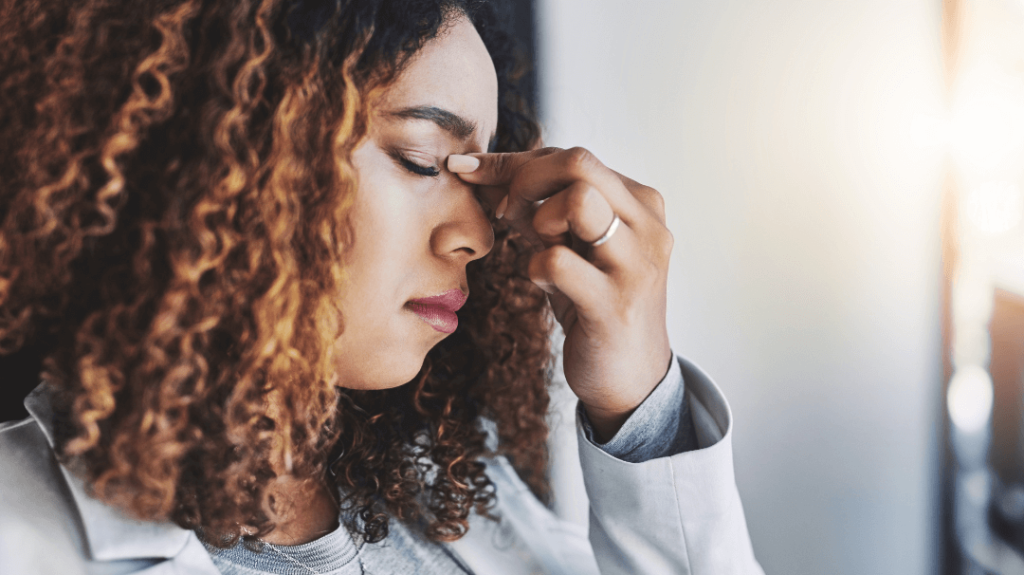 A woman with curly hair looking stressed and overwhelmed, pressing her fingers to the bridge of her nose with her eyes closed.