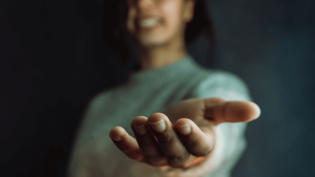 A close-up of an open hand reaching forward with a smiling woman softly blurred in the background, symbolizing support and connection.