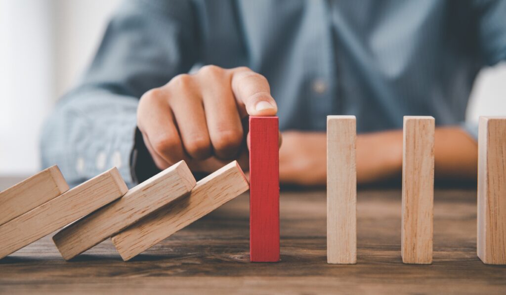 A close-up, selective-focus photograph of a person's hand placing an index finger on top of a single, upright bright red wooden domino block. To the left, a row of natural-colored wooden blocks has already toppled over in a chain reaction that was stopped by the red block. To the right, the remaining sequence of natural wooden blocks stands completely upright and unaffected.