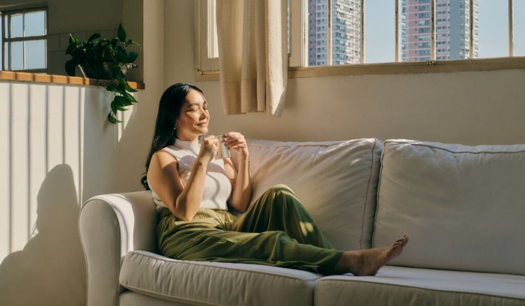 A woman with long black hair tied back is relaxing on a white fabric sofa, smiling with her eyes closed while holding a small ceramic mug. She is wearing a white sleeveless top and olive green trousers. Warm, bright sunlight streams through a window behind her, casting long shadows on the wall. The background shows an urban view with high-rise buildings outside the window.