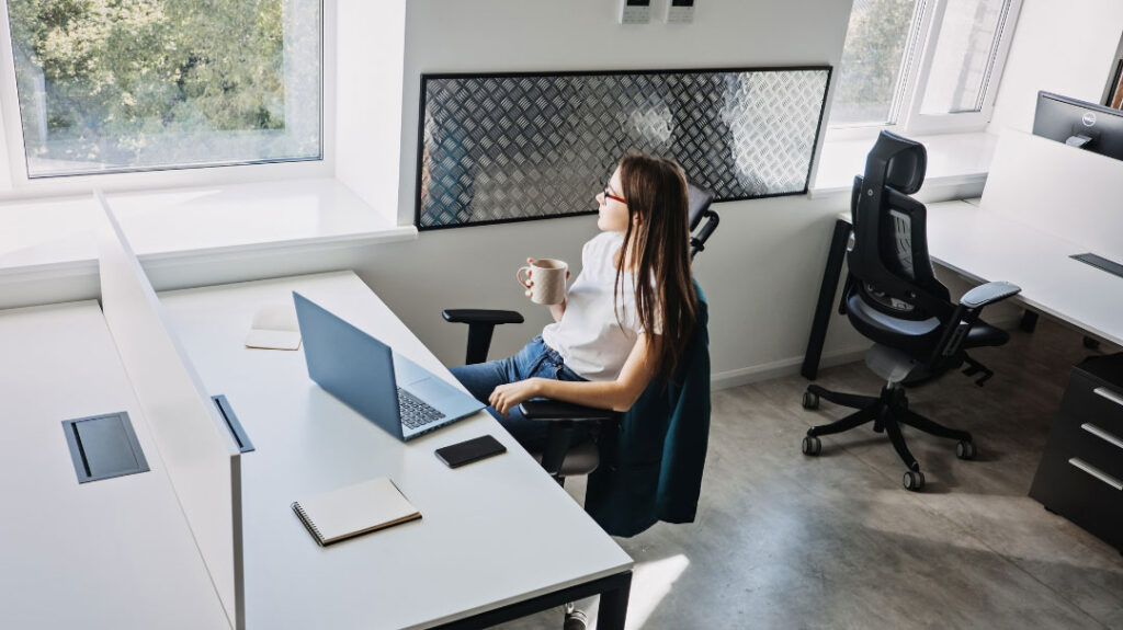A woman with glasses sitting at a modern white office desk in a bright, minimalist workspace, holding a coffee mug and looking out the window. Her laptop, a smartphone, and a notebook rest on the desk in front of her. The surrounding office area features empty desks, ergonomic chairs, and a clean, professional aesthetic.