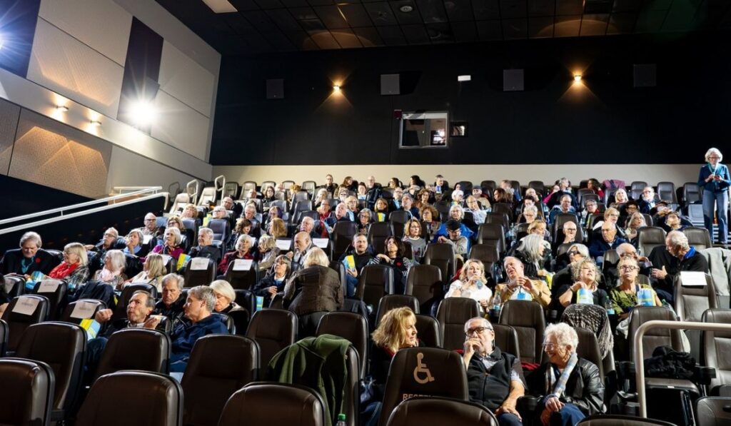 A large audience seated in a dark, auditorium-style theater, watching a presentation or film.