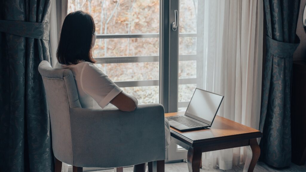 A woman sitting in a chair, looking out a window at a blurred, outdoor seasonal scene, with a laptop open on the table beside her.
