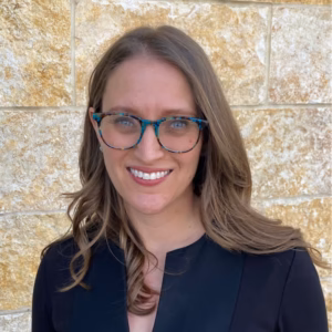 A professional headshot of a smiling woman with long, light-brown hair and glasses with a colourful, patterned frame. She is wearing a simple black top, set against a light-coloured, textured stone-wall background.