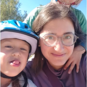 A smiling woman with glasses and long dark hair, wearing a blue bicycle helmet. She is holding a small child who is wearing a helmet and making a playful face at the camera. The background shows bright, sunny outdoor greenery.