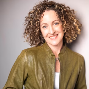 A professional headshot of a smiling woman with shoulder-length, wavy, dark-brown hair. She is wearing a dark-olive leather jacket over a white top and a delicate silver necklace, set against a soft, neutral grey background.