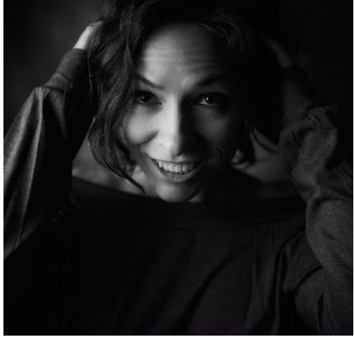 A black-and-white, close-up portrait of a woman smiling broadly at the camera. She has her hands raised to her head, with her fingers touching her hair. The lighting highlights her face, creating a playful and intimate mood.