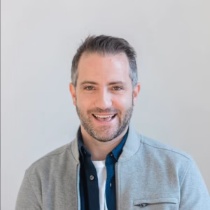 A professional headshot of a smiling man with short, dark-brown hair and a trimmed beard. He is wearing a dark-collared shirt under a light-grey bomber-style jacket, set against a soft, neutral off-white studio background.