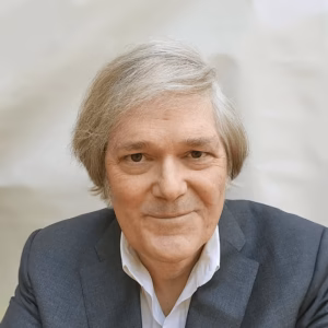 A professional headshot of a smiling man with thick, wavy grey hair. He is wearing a dark grey suit jacket over a white collared shirt, set against a soft, neutral off-white studio background.