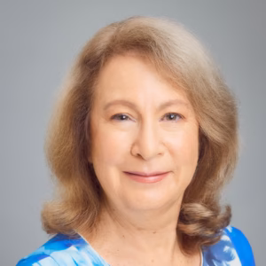 A professional headshot of a smiling woman with shoulder-length, soft-wavy, light-brown hair. She is wearing a bright blue top with a subtle pattern, set against a soft, neutral, light-grey studio background.