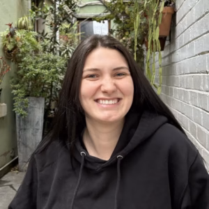 A close-up headshot of a smiling young woman with long, straight dark hair. She is wearing a black hooded sweatshirt, set against an outdoor background of a brick wall and green potted plants.