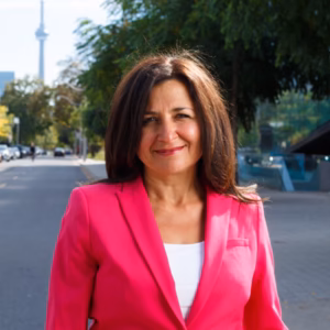 A professional headshot of a smiling woman with shoulder-length, wavy dark brown hair. She is wearing a bright pink blazer over a white shirt, set against a soft, outdoor urban background with trees and a blurry cityscape.