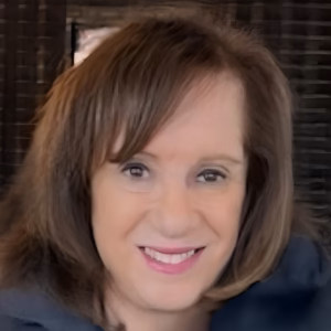 A professional headshot of a smiling woman with shoulder-length, wavy brown hair, facing the camera. She is wearing a dark, casual top, set against a dark, textured stone background.