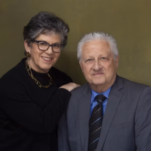 A professional portrait of a smiling older couple, David and Deborah Cooper. Deborah has short, dark hair and is wearing a black top with a gold chain necklace. David has grey hair and is wearing a grey suit jacket, a white collared shirt, and a dark striped tie. They are posed together against a soft, textured olive-green background.
