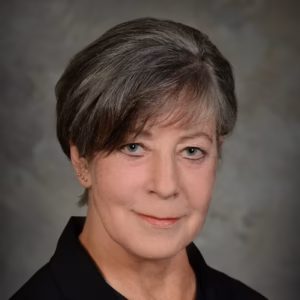 A professional headshot of a smiling woman with short, textured brown-and-grey hair. She is wearing a simple black top, set against a soft, neutral, mottled grey studio background.