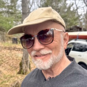 A close-up portrait of a smiling man with a white beard and glasses, wearing a beige baseball cap. The background is a soft, out-of-focus view of trees and the back of a car, creating a casual, outdoor atmosphere.