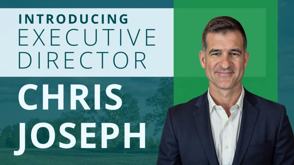 A promotional graphic for a blog post announcing the appointment of Chris Joseph as Executive Director. The image features a professional portrait of Chris Joseph, a man with short brown hair wearing a suit, set against a green, tree-filled background, with the text "Introducing Executive Director Chris Joseph" clearly displayed.