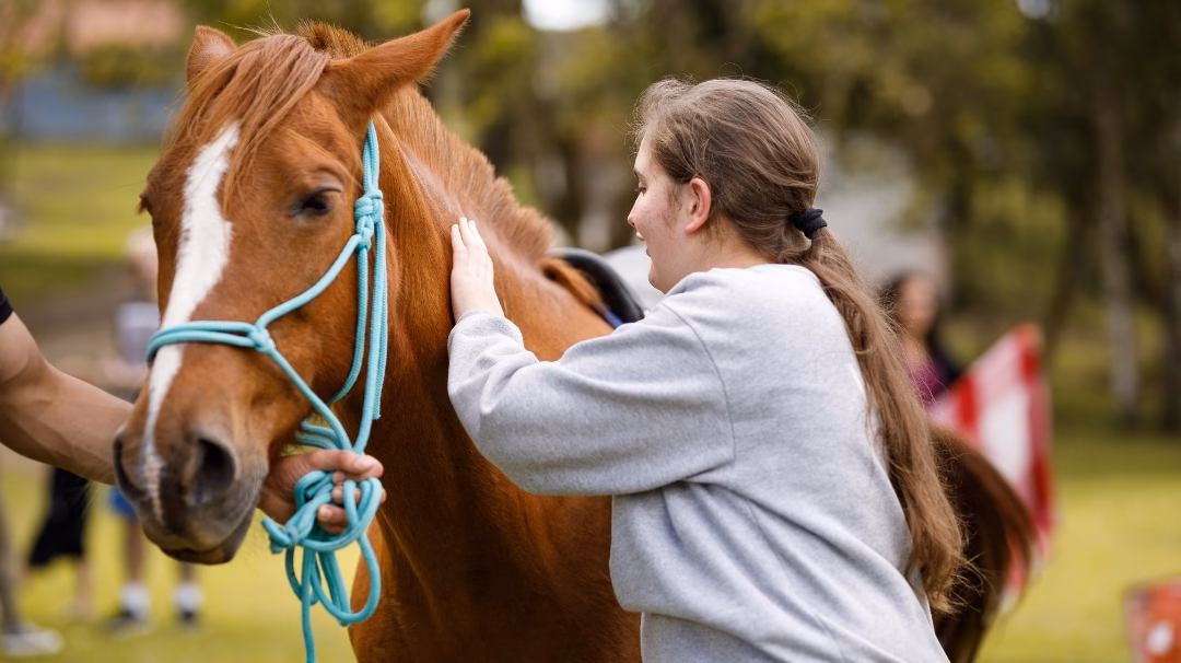 Animal and Nature Therapy — A young adult woman with a horse, green grass and trees in the background with some people out of focus in the distance, for animal and therapy at Eli's Place